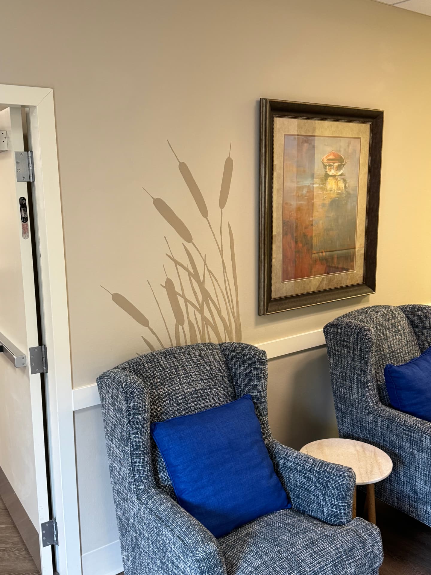 Living room at Lighthouse Memory Care with two blue tweed armchairs, a painted cattail mural and a framed painting