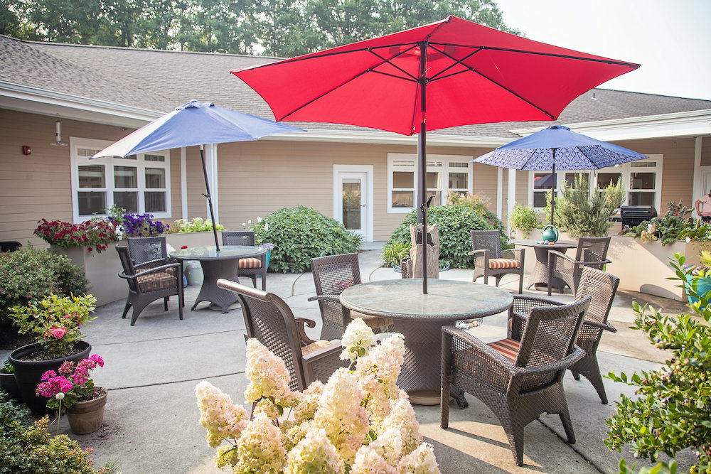Wide view of the courtyard at Lighthouse Memory Care with a large red umbrella over a glass topped table, a blue umbrella and a patterned umbrella over further seating groups, and white hydrangeas in front